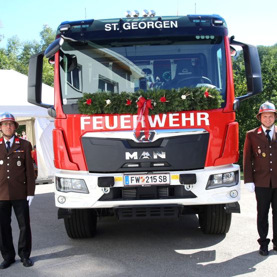 Zwei Feuerwehrleute stehen neben einem roten MAN-Feuerwehrfahrzeug mit 'FEUERWEHR' und 'ST. GEORG' Text, geschmückt mit einer Weihnachtsgirlande.