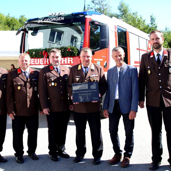 Fünf Männer in Uniform stehen vor einem Feuerwehrauto und halten eine Plakette. Ein Mann trägt einen blauen Anzug.