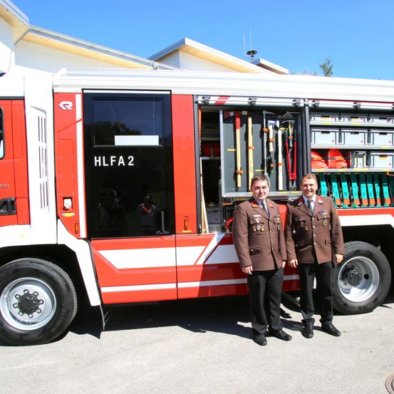 Zwei Feuerwehrleute in Uniform stehen vor einem Feuerwehrwagen mit der Aufschrift 'HLFa2'. Der Wagen ist geöffnet und zeigt verschiedene Feuerwehrgeräte und Ausrüstung.