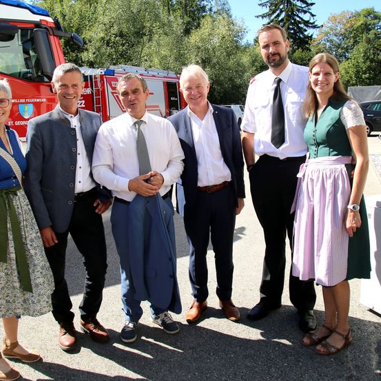 Fünf Personen, darunter drei Männer und zwei Frauen, stehen vor einem roten Feuerwehrauto. Sie sind alle formell gekleidet, wobei der Mann ganz rechts ein traditionelles Outfit trägt.