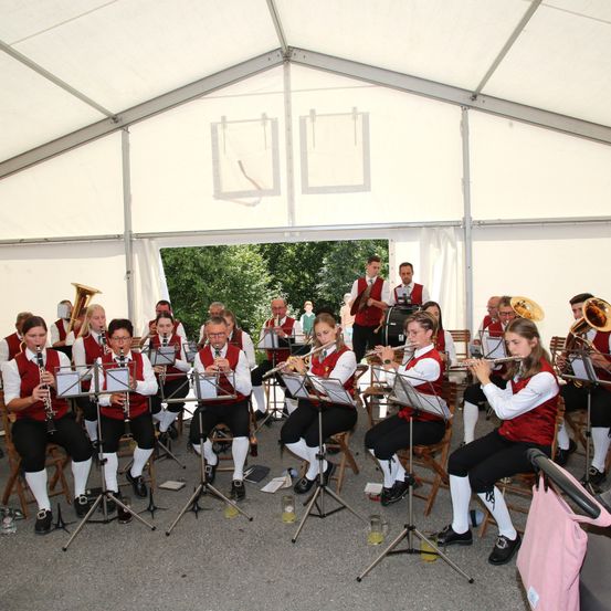 Ein Blasorchester spielt in einem Zelt mit weißem Dach, wobei Musiker verschiedene Instrumente unter der Aufsicht von zwei Männern spielen.