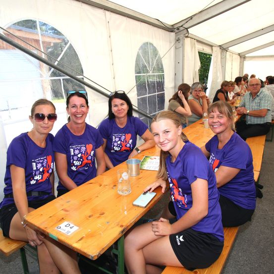 Fünf Frauen in passenden lila T-Shirts sitzen an einem Tisch in einem Zelt und lächeln für die Kamera. Ein junges Mädchen sitzt neben ihnen, und mehrere Personen sitzen an anderen Tischen im Hintergrund.