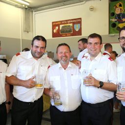 Vier Männer in Uniform, lächelnd und Biergläser haltend, stehen zusammen in einem Raum mit einem Banner im Hintergrund.
