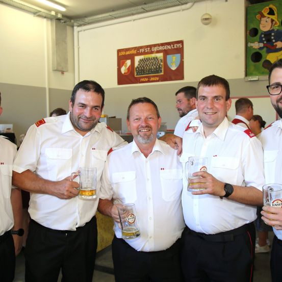 Vier Männer in Uniform, lächelnd und Biergläser haltend, stehen zusammen in einem Raum mit einem Banner im Hintergrund.