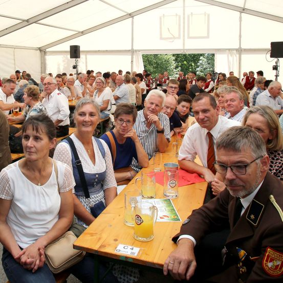 Eine Gruppe von Menschen sitzt an einem langen Tisch in einem Zelt, einige mit Getränken und andere in Gesprächen vertieft. Die Umgebung scheint eine soziale Zusammenkunft oder Veranstaltung zu sein.
