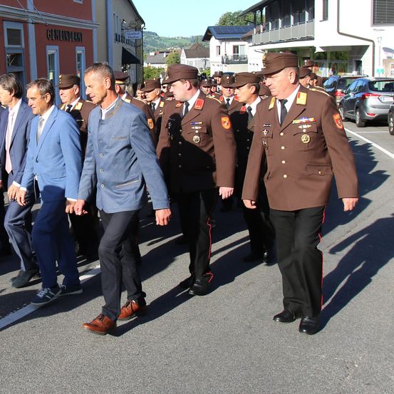Eine Gruppe von Männern in Uniformen und Anzügen, die auf einer Straße gehen, möglicherweise bei einer zeremoniellen Prozession.