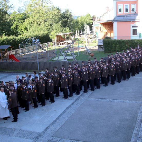 Eine Gruppe uniformierter Personen, möglicherweise Militärangehörige, steht in Formation vor einem Gebäude mit einem Spielplatz im Hintergrund.