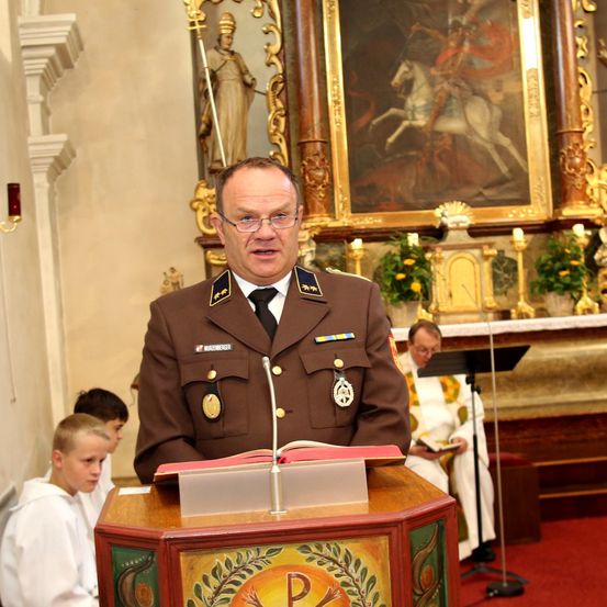 Ein Militäroffizier steht an einem Podium, liest aus einem Buch, mit kirchlichem Hintergrund und sitzendem Klerus.