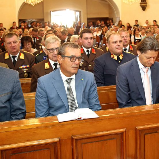 Eine Gruppe von Männern in Anzügen und Krawatten sitzt auf einer Kirchenbank, einige tragen Militäruniformen. Ein offenes Buch liegt vor ihnen auf der Bank.