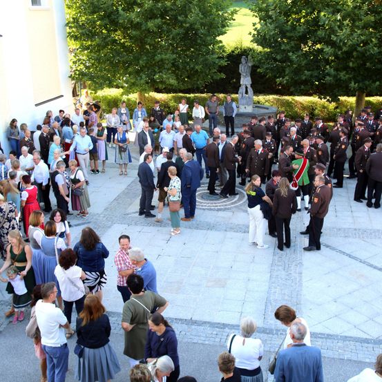 Eine große Gruppe von Menschen ist in einem Außenbereich versammelt, möglicherweise für eine formelle Veranstaltung. Einige tragen Uniformen, und eine Statue ist im Hintergrund zu sehen.