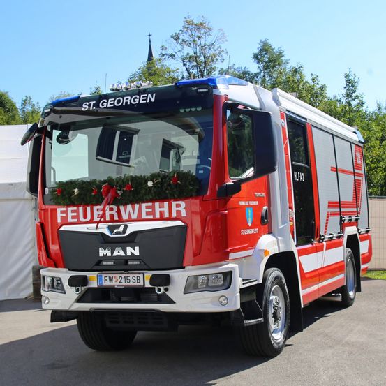 Ein rot-weißer MAN Feuerwehrwagen mit 'Feuerwehr' und 'St. Georg' Beschriftung, geparkt auf einem Parkplatz.