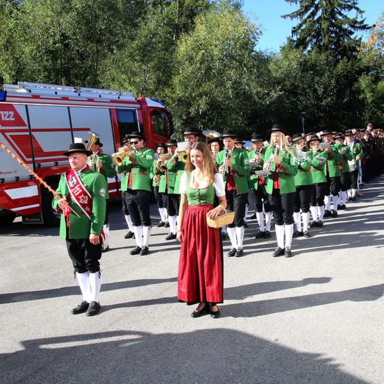 Eine Blaskapelle in traditionellen grünen Uniformen, angeführt von einer Frau in einem roten Kleid, tritt vor einem Feuerwehrauto auf.