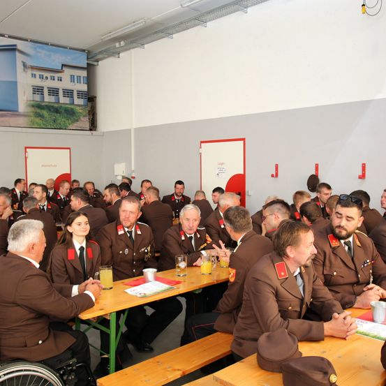 Eine Gruppe uniformierter Personen, wahrscheinlich Militärangehörige, sitzen an Tischen in einem Saal und führen Gespräche und trinken Getränke.
