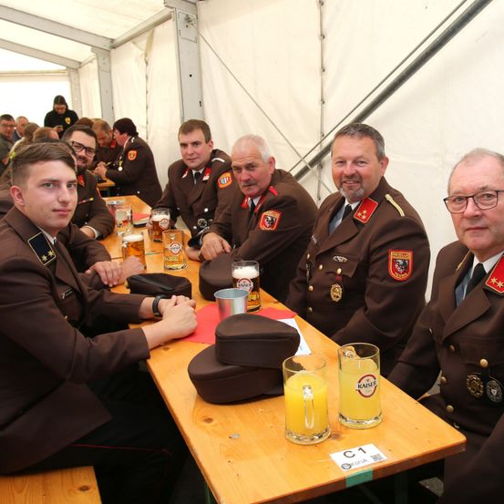 Eine Gruppe von Männern in Militäruniformen sitzt an einem Tisch in einem Zelt, lächelt und posiert für ein Foto. Sie haben Gläser Bier und eine Tasse Orangensaft auf dem Tisch.