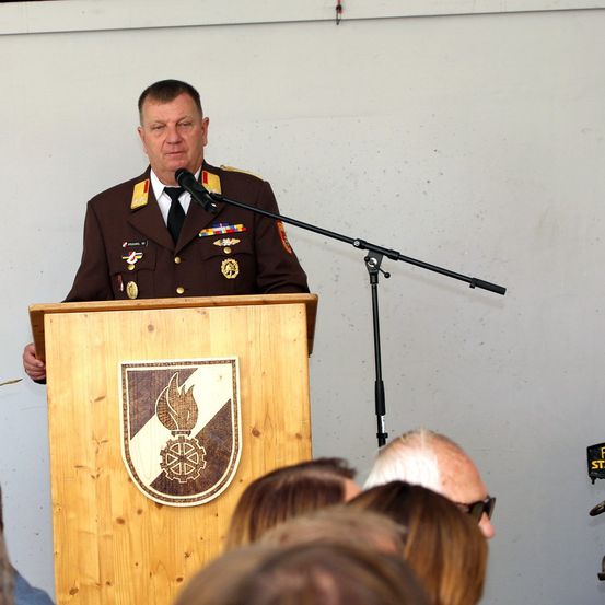 Ein Militäroffizier in voller Uniform steht an einem Rednerpult und spricht vor einem Publikum. Das Pult hat ein hölzerne Design mit einem Wappenschild. Ein Mikrofon steht vor ihm.