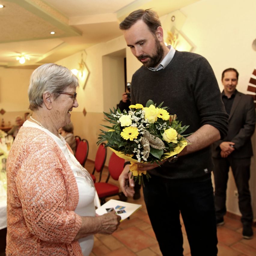 Ein Mann mit einem Strauß gelber Blumen steht lächelnd vor einer älteren Frau, während zwei Männer in Anzügen von hinten zusehen.
