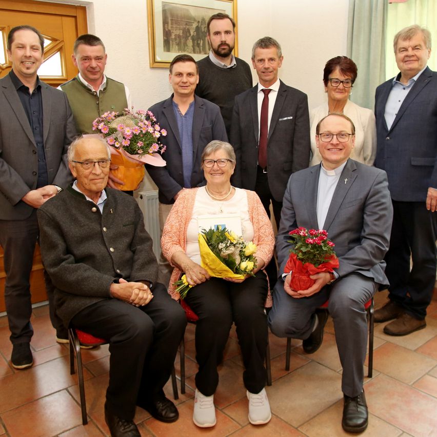 Eine Gruppe von Menschen posiert für ein Foto. Einige von ihnen halten Blumen. Eine Frau sitzt auf einem Stuhl und ein Mann steht hinter ihr. Ein Mann trägt eine Krawatte und eine Frau trägt eine Brille. Ein Bilderrahmen ist an der Wand hinter ihnen angebracht.