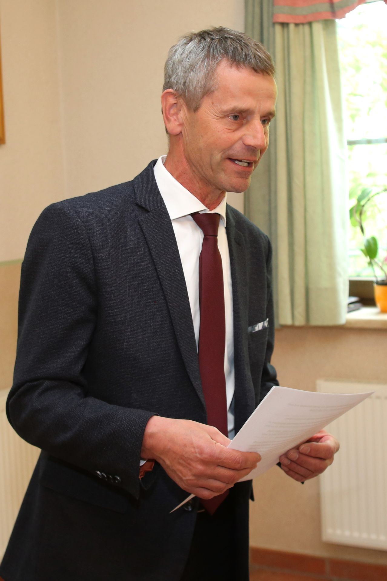 Ein Mann steht und lächelt, während er ein Blatt Papier in den Händen hält. Er trägt einen dunklen Anzug, ein weißes Hemd und eine dunkelrote Krawatte.