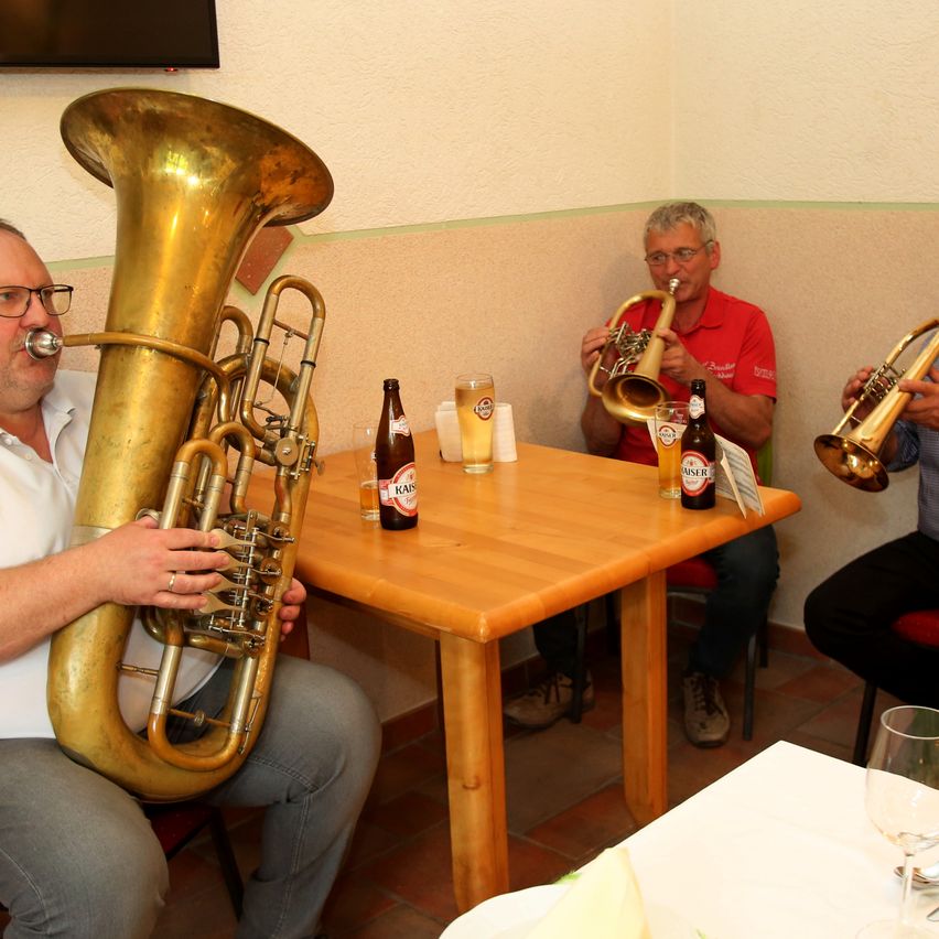 Drei Männer in einem Raum, die Trompeten spielen. Einer sitzt mit einem Tuba. Flaschen und Gläser auf dem Tisch.