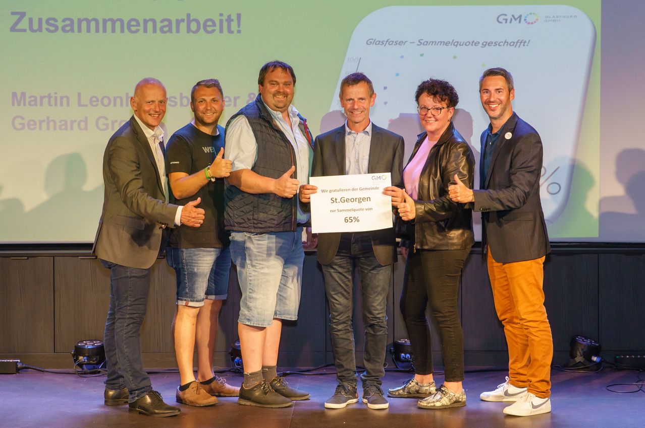 Fünf Männer und eine Frau stehen auf der Bühne und halten ein Zertifikat. Die Frau trägt eine Lederjacke und eine Brille. Auf dem Zertifikat steht 'GMO' und 'St. Georg' mit einem Prozentsatz. Sie alle zeigen den Daumen nach oben.