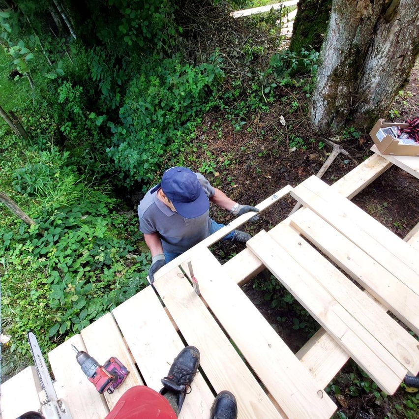 Ein Mann mit blauer Mütze und Handschuhen arbeitet an einer Holzterrasse im Wald, mit Werkzeug und einer Kiste in der Nähe.