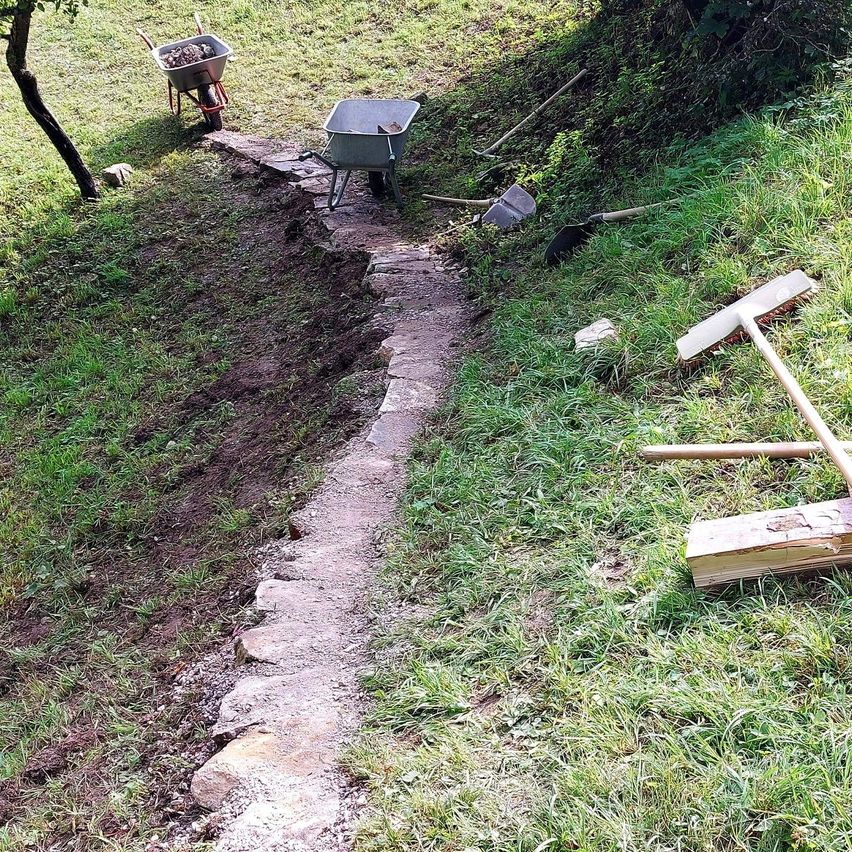 Ein Grasbereich mit einem Steinpfad und verstreuten Werkzeugen. Zwei Schubkarren, einer beladen, stehen auf beiden Seiten. Verschiedene Gartenwerkzeuge liegen auf dem Boden.