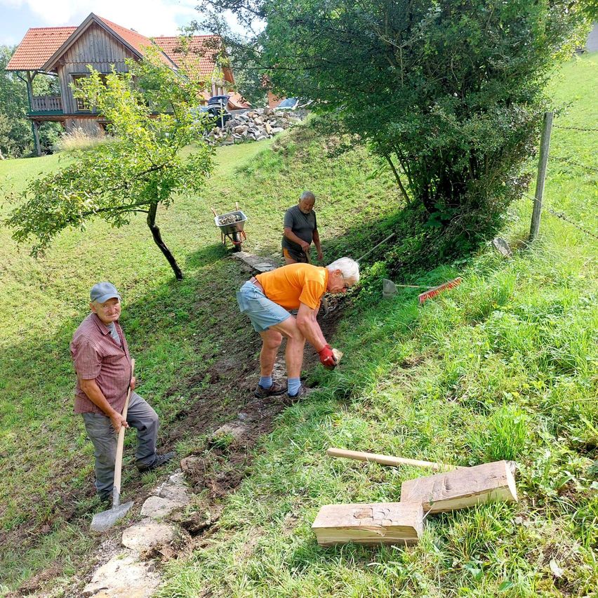 Drei Männer arbeiten auf einem grün bewachsenen Hang, jeder mit einer Schaufel. Ein Mann in orangefarbener und blauer Shorts beugt sich vor, während ein anderer Mann mit einem Schubkarren steht. Im Hintergrund befindet sich ein Haus mit rotem Dach und einem Haufen Steine.
