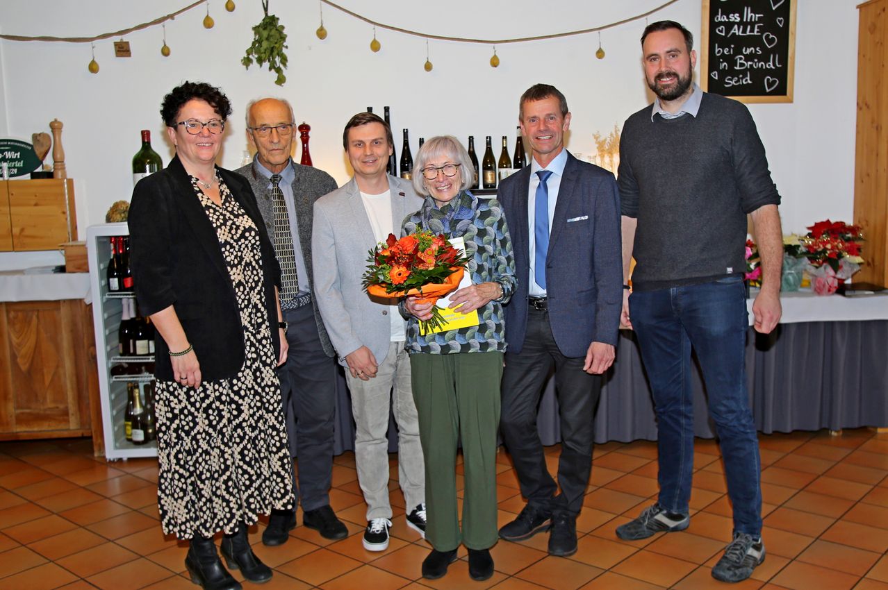 Eine Gruppe von Menschen steht zusammen mit einer Frau, die einen Blumenstrauß hält. Sie scheinen sich bei einer Veranstaltung oder Feier zu befinden.