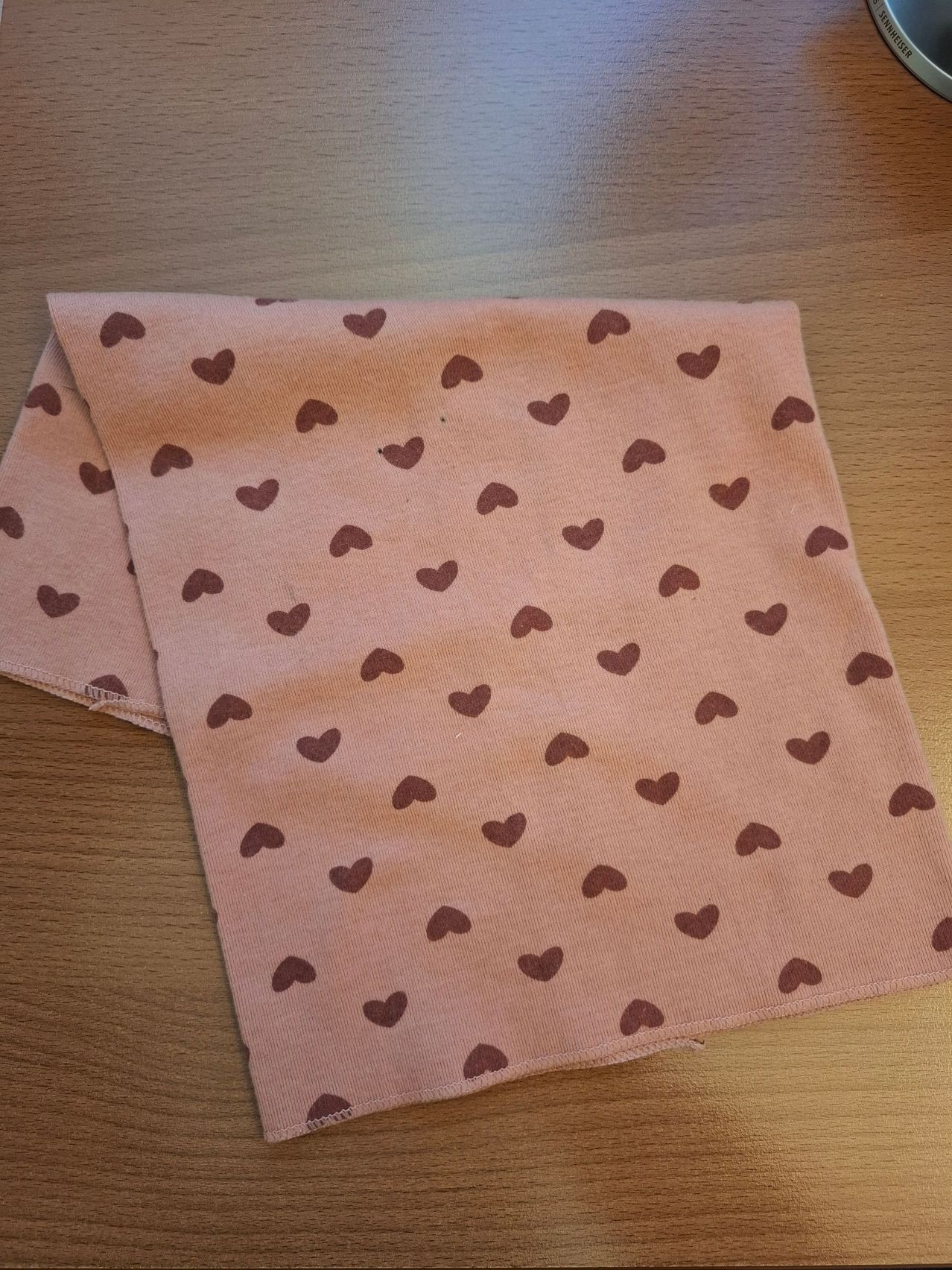 Ein hellrosa Stoff mit braunen Herzmustern auf einer Holzoberfläche. Der Stoff ist gefaltet und scheint ein Taschentuch zu sein.