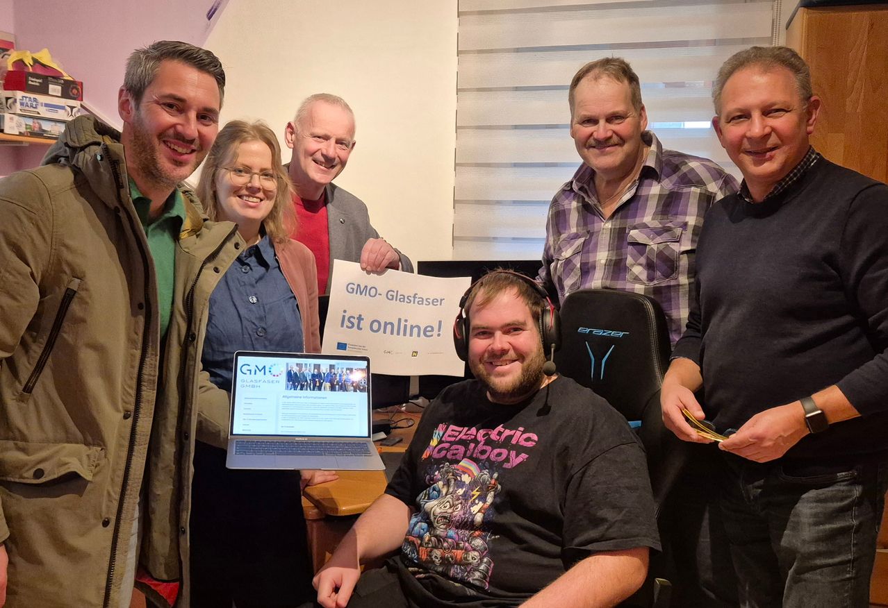 Eine Gruppe von Menschen steht um einen Laptop herum. Einer hält ein Schild mit der Aufschrift GMO-Glasfaser ist online. Sie alle lächeln.