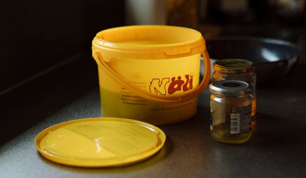 Ein gelber Eimer mit der Aufschrift 'Nöll' steht mit offenem Deckel auf einer Arbeitsplatte, begleitet von zwei Gläsern, eines mit Etikett und das andere mit Metalldeckel.