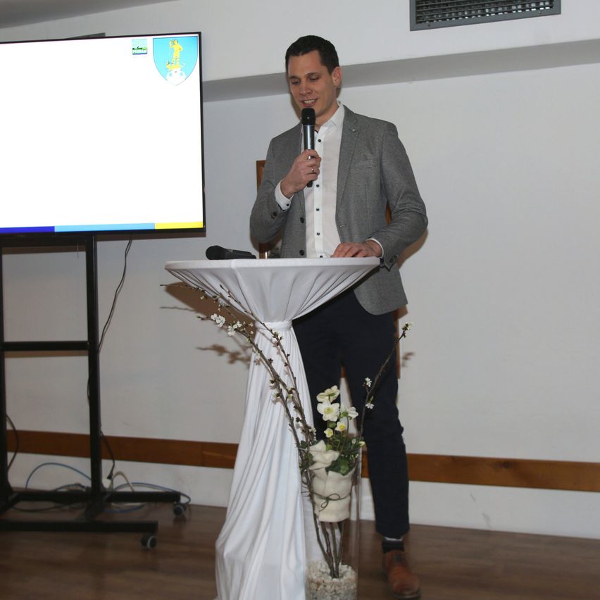 Ein Mann steht an einem Rednerpult, hält ein Mikrofon und spricht. Hinter ihm befindet sich ein Bildschirm, der eine Präsentation zeigt. Das Pult ist mit einem weißen Tuch und einer Blumendekoration geschmückt. Die Umgebung ist in einem Innenraum mit Holzboden.