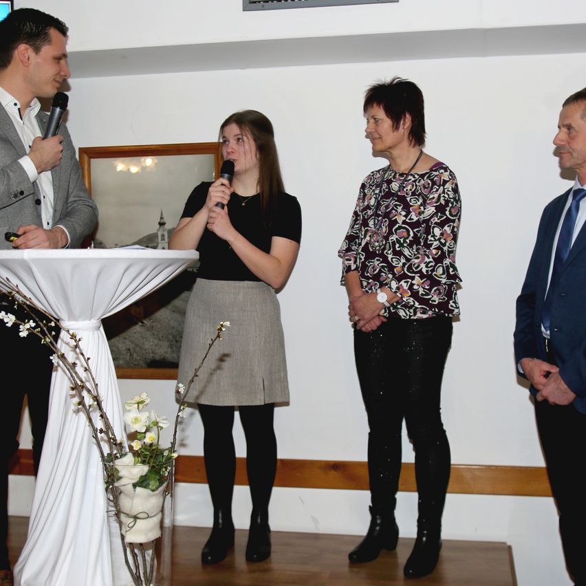 Eine Frau spricht in ein Mikrofon, während sie vor einem Tisch mit Blumen steht, wobei zwei Männer und eine weitere Frau zuhören. Sie befinden sich in einem Raum mit weißen Wänden.