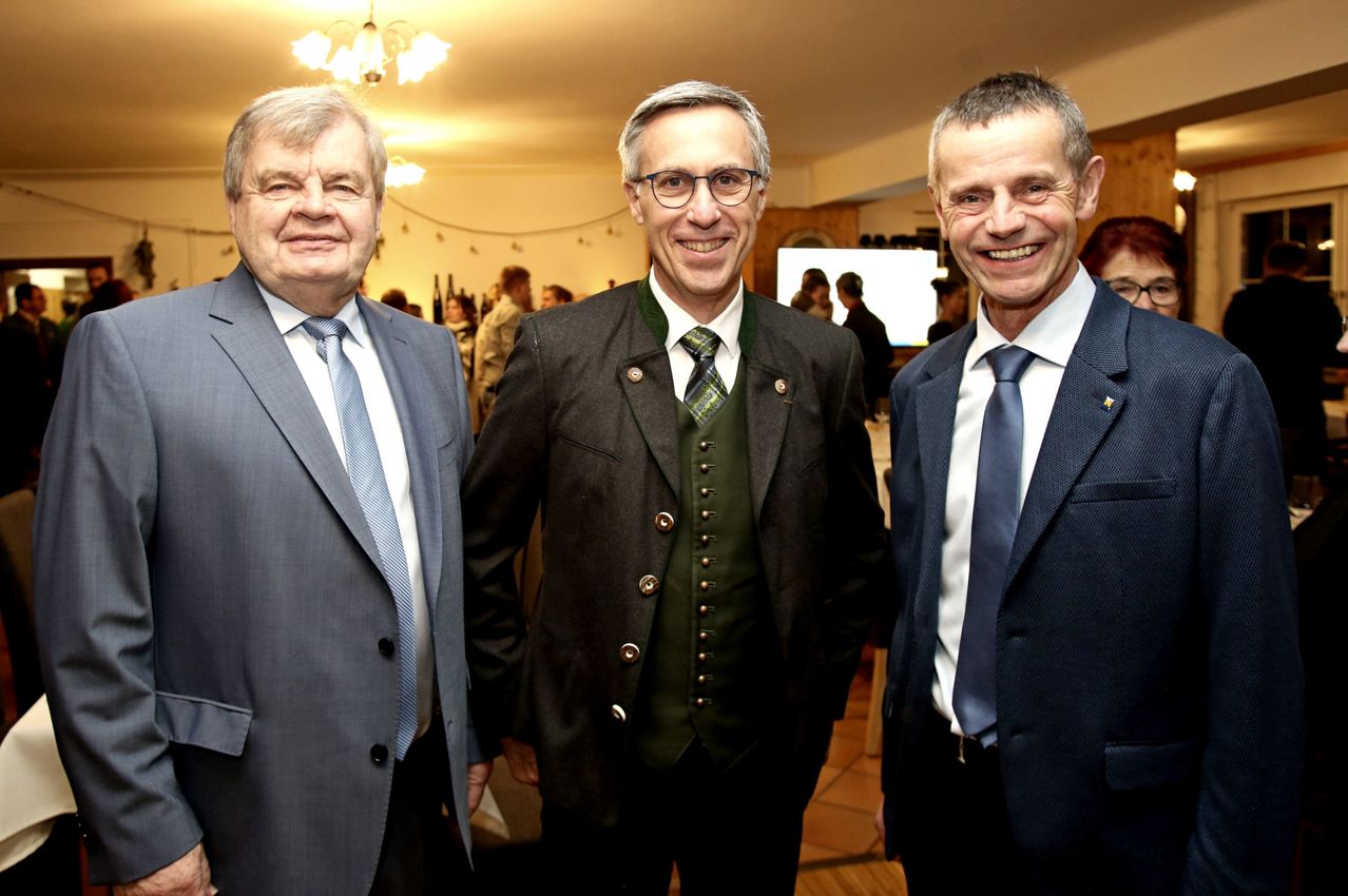 Drei Männer in Anzügen und Krawatten lächeln und posieren für ein Foto. Der Mann in der Mitte trägt eine grüne Weste mit goldenen Knöpfen. Hinter ihnen ist ein Raum mit einer Kronleuchter und einer Projektionsleinwand zu sehen.