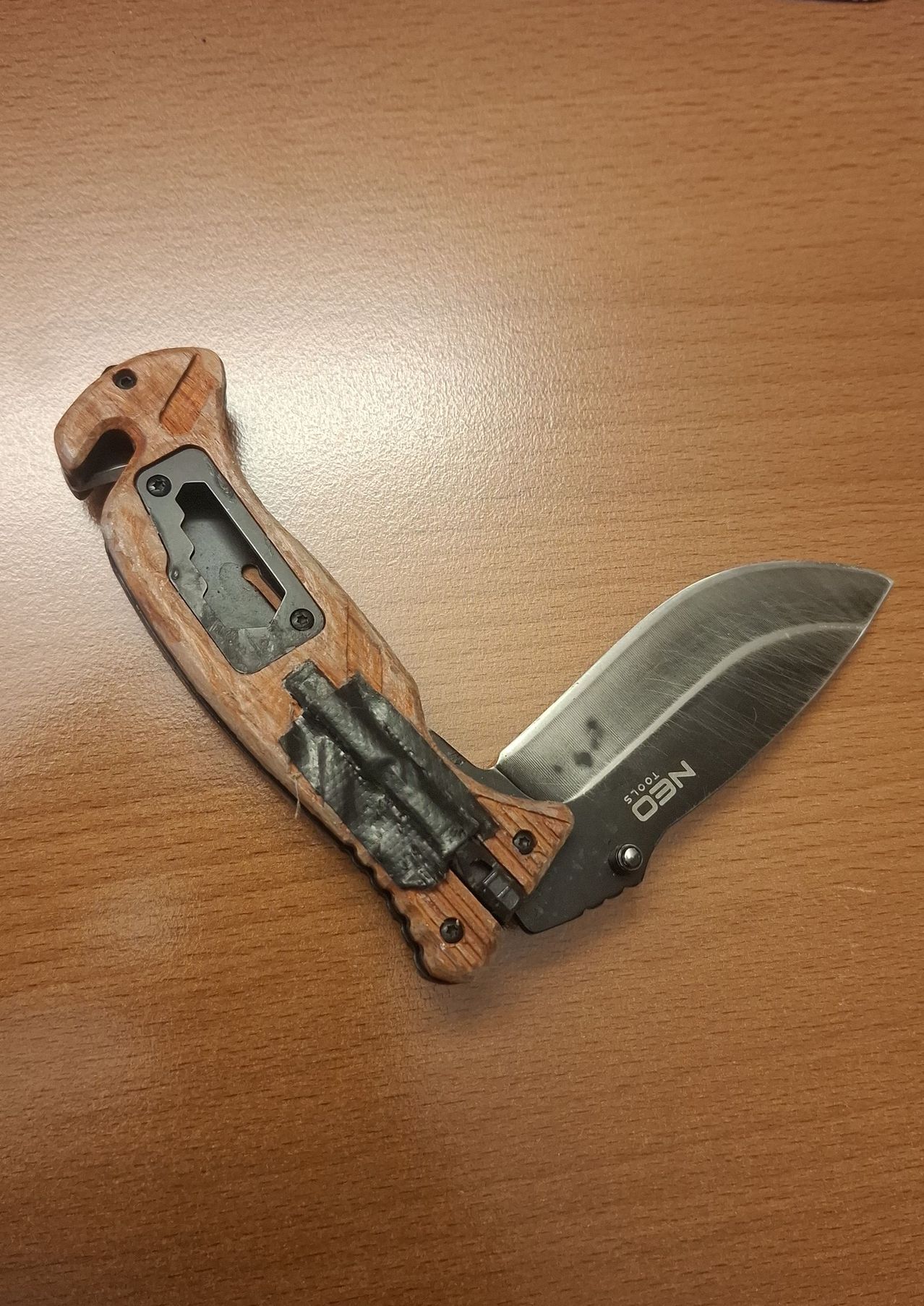 Ein Nahaufnahme eines Taschenmessers mit einem Holzgriff, auf einer Holzoberfläche platziert. Die Klinge ist offen und schwarzer Klebeband ist um den Griff gewickelt.