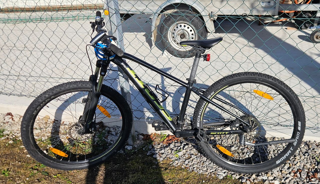 Ein schwarzes Scott Mountainbike steht auf Kies und lehnt an einem Maschendrahtzaun. Im Hintergrund ist ein Fahrzeug mit einem großen Rad zu sehen.