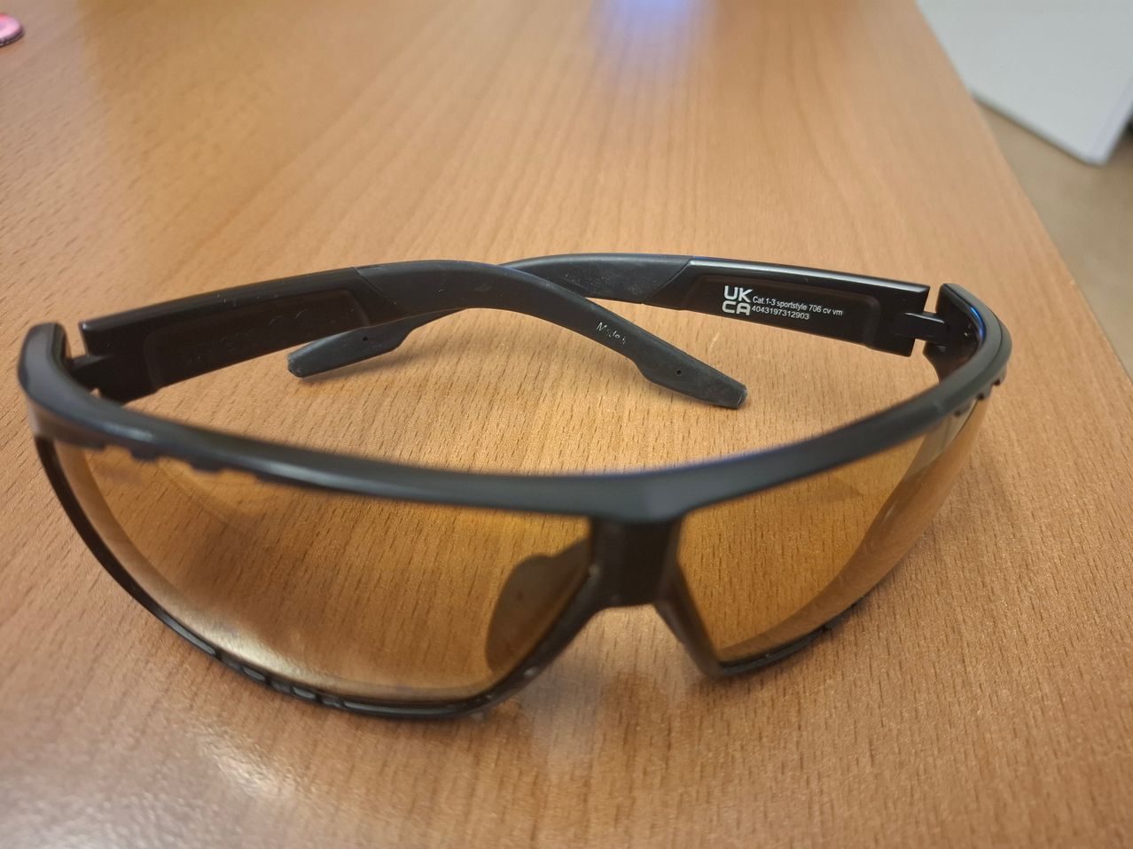 Schwarze Schutzbrille mit braunen Gläsern, beschriftet UK CR, auf einer Holzoberfläche platziert.