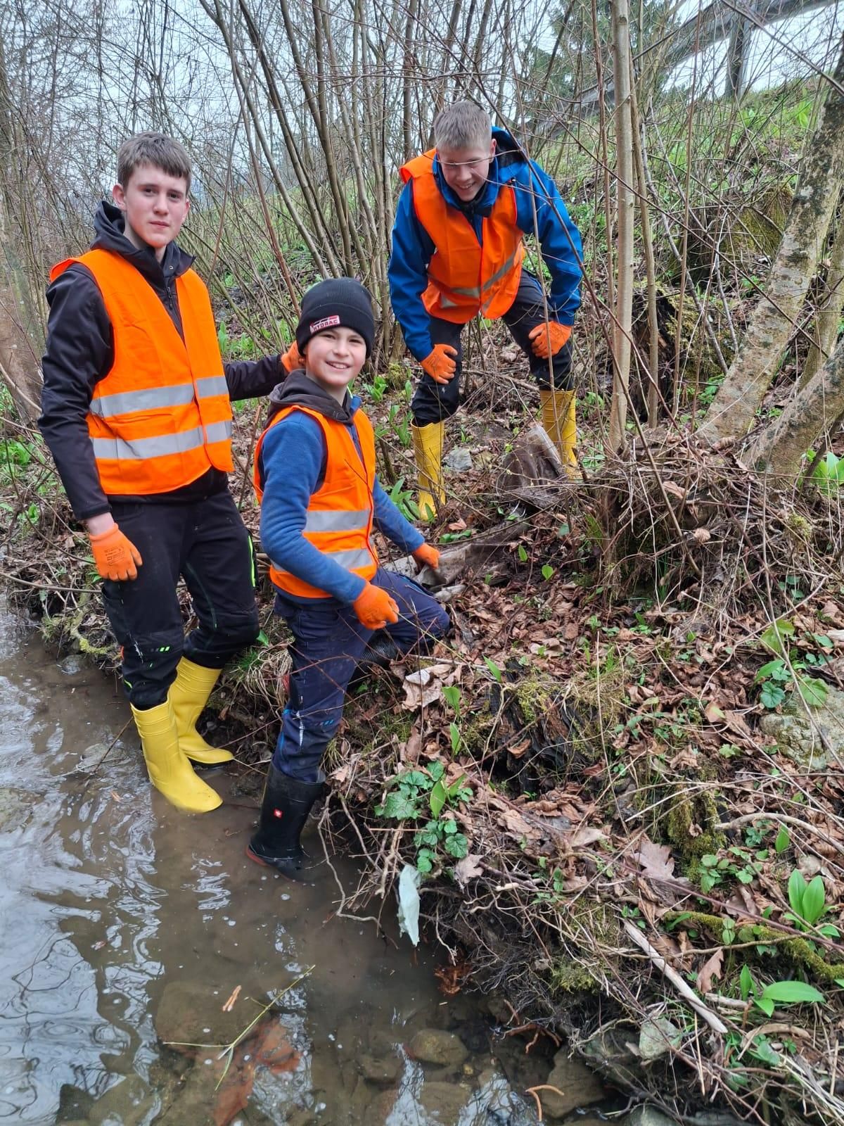 Three young individuals in high visibility vests and boots work in a muddy area, one holding a tool.