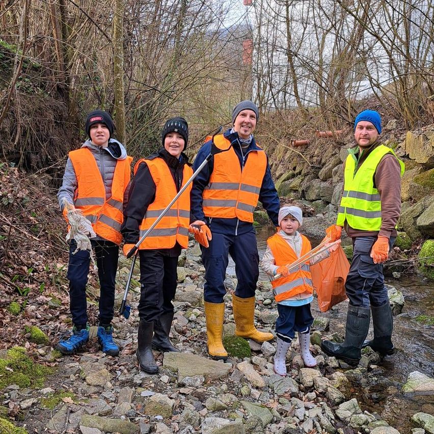 Five people in safety vests are cleaning a riverbank. One holds a net, another a trash bag. The area is rocky with trees and moss.