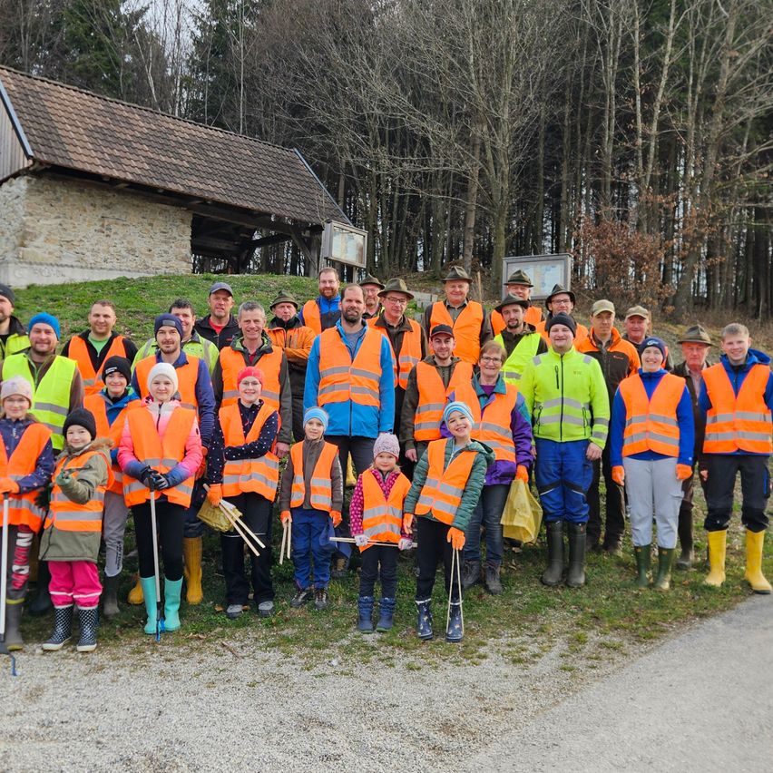 A group of people in orange vests stands in a line in front of a building with a forest in the background.