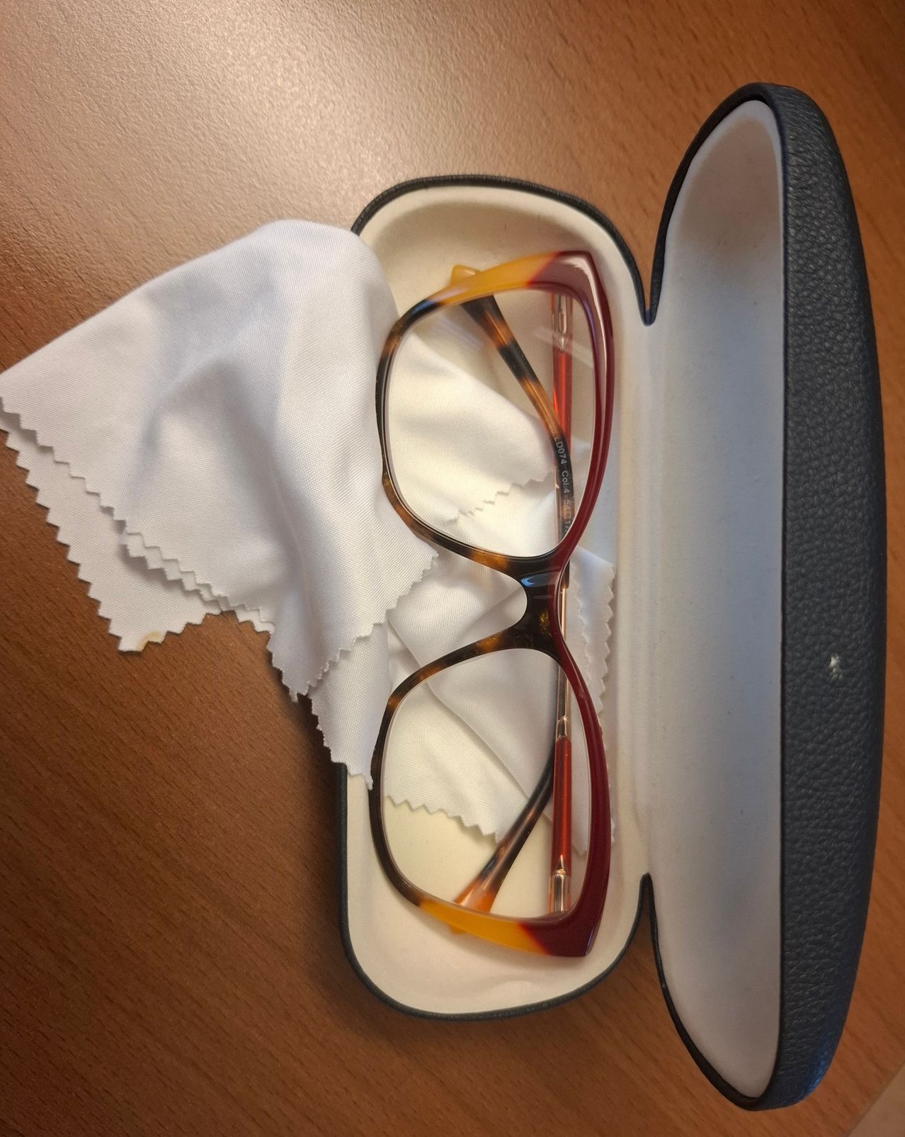 Ein Paar roter Brille liegt in einem schwarzen Etui. Ein weißer Stoff ist gefaltet im Etui. Der Tisch hat eine hölzerne Oberfläche.