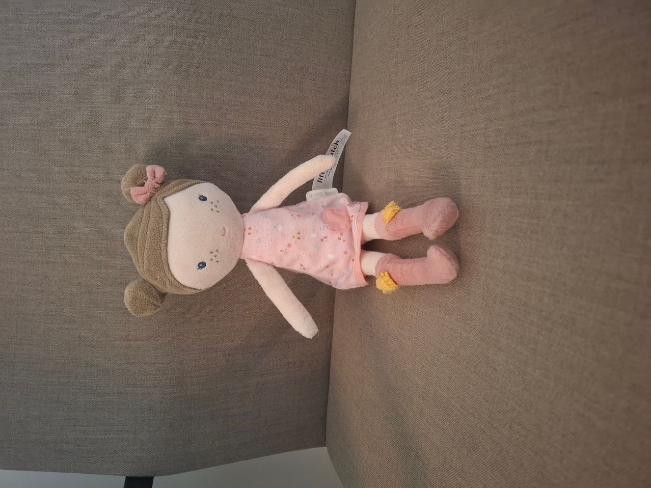Eine Plüschpuppe in einem rosa Kleid und rosa Socken mit einem Schild am linken Arm liegt auf einer braunen Couch.