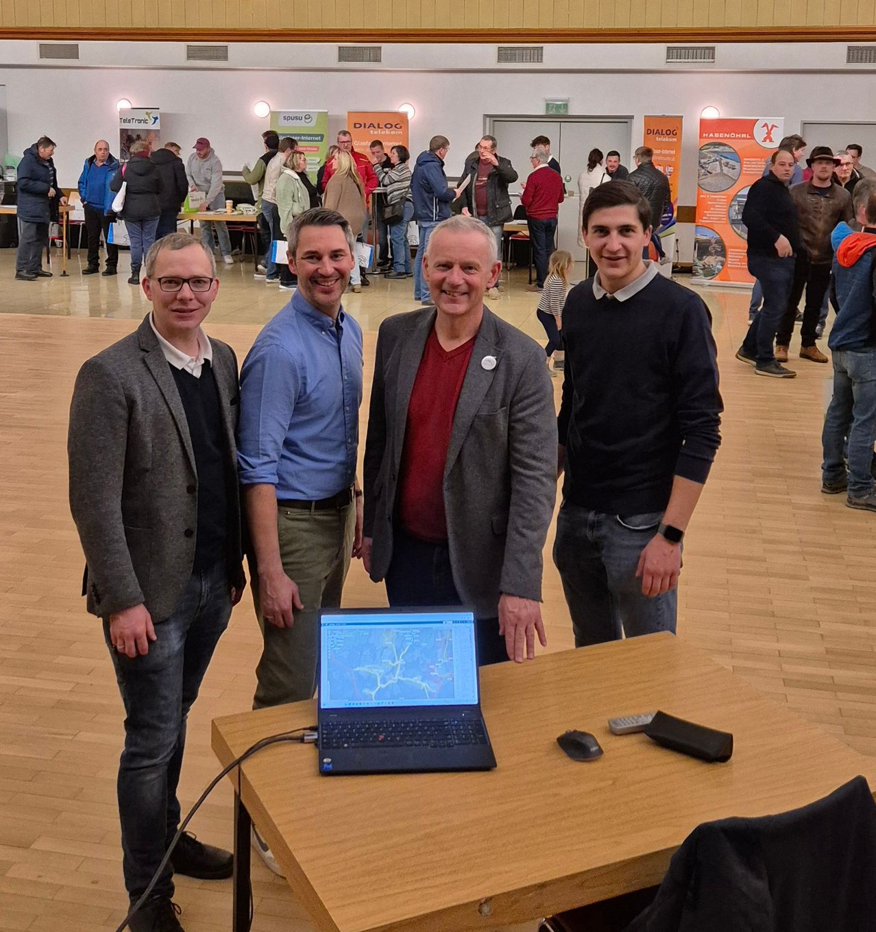 Vier Männer stehen neben einem Tisch mit einem Laptop und lächeln in einer Innenumgebung mit Holzboden und verschiedenen Menschen im Hintergrund.