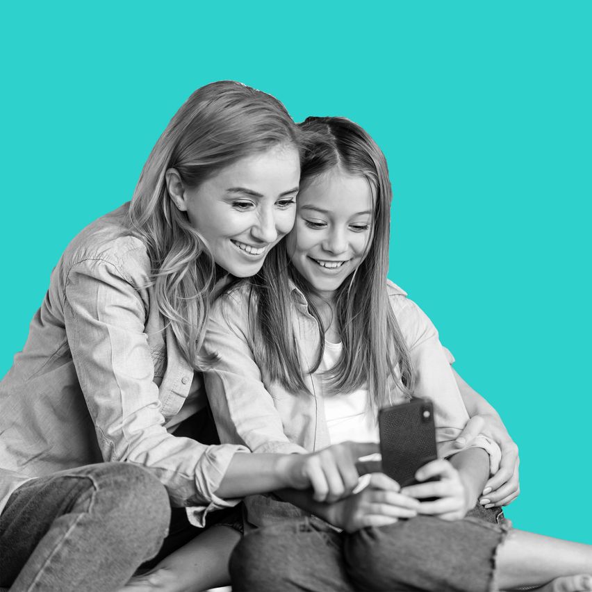 Zwei Mädchen sitzen auf einem grünen Hintergrund, lächelnd, eines hält ein Telefon, während das andere auf den Bildschirm schaut.