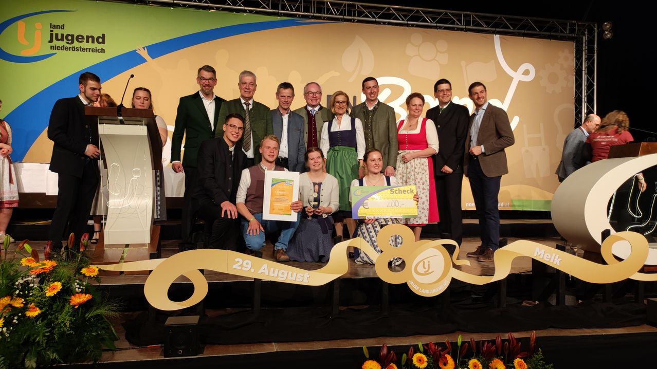 Eine Gruppe von Menschen auf der Bühne, lächelnd, mit Urkunden und einem großen Scheck in der Hand, mit einem Banner im Hintergrund und Blumen im Vordergrund.