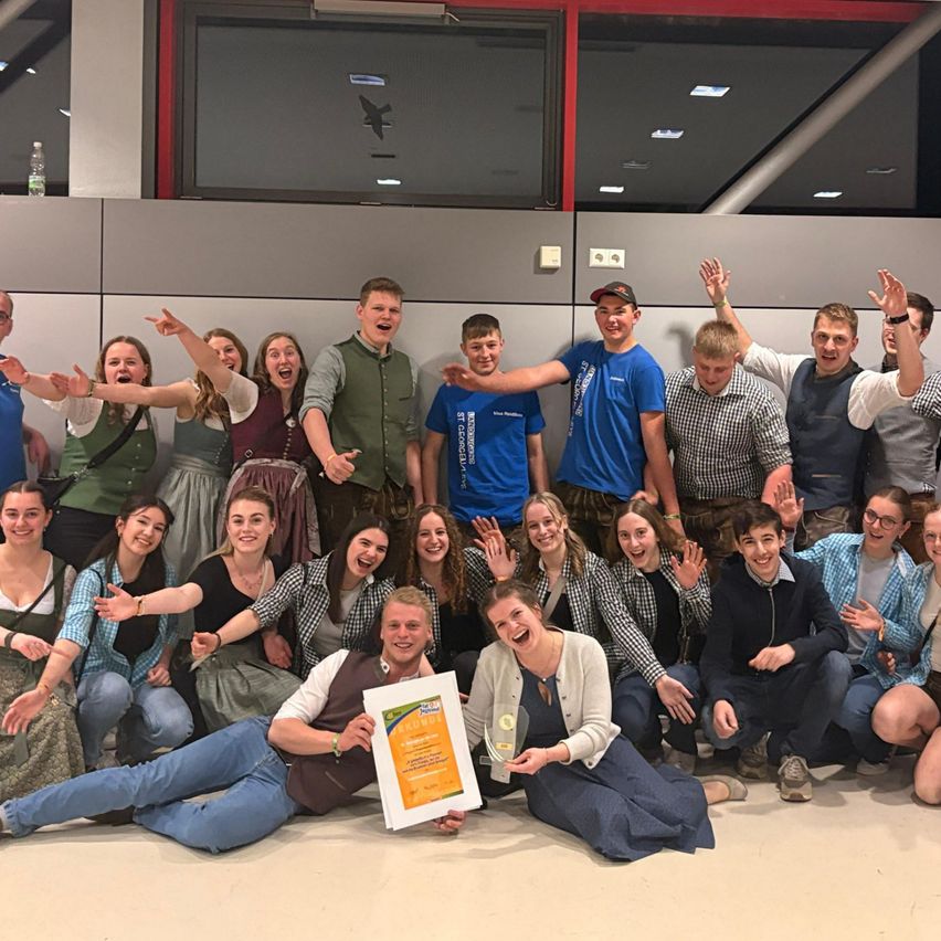 Eine Gruppe von Menschen posiert für ein Foto mit einem gerahmten Zertifikat in einem Raum mit einer grauen Wand und Glasfenstern.