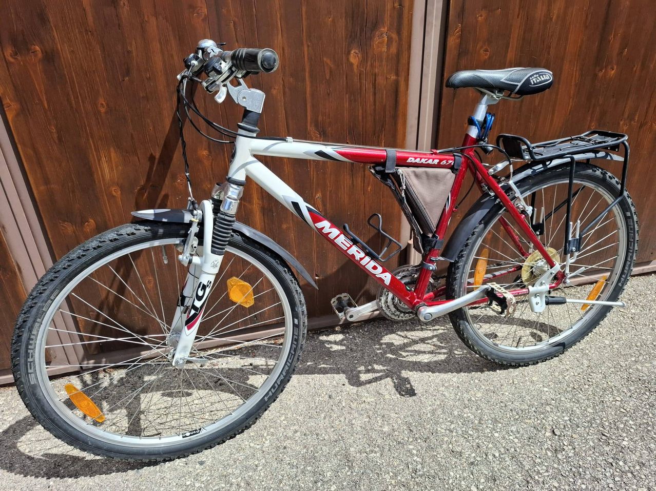 A red and white Merida bicycle is parked against a wooden wall. The bike has a black seat, silver handlebars, and a black and silver rear rack.