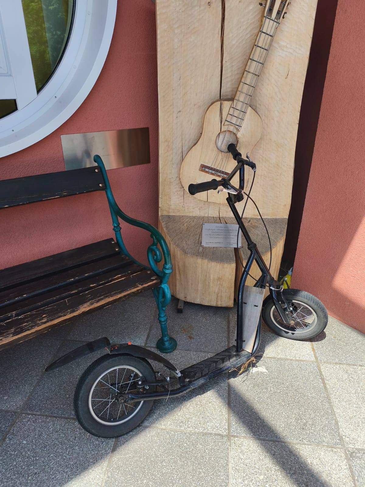 Ein schwarzer Roller steht neben einer Bank und einer hölzernen Gitarrenskulptur an einer rosa Wand.