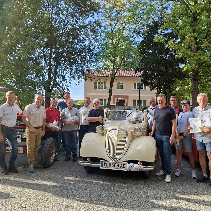 A group of men stands around an old car, holding gifts and smiling for the camera. Behind them is a building with a red roof and a tractor.