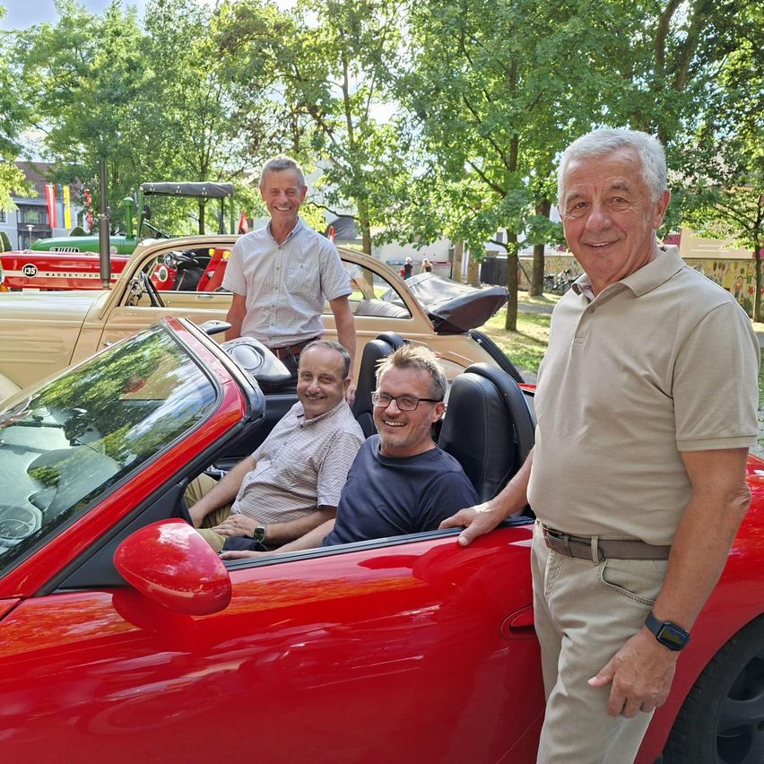 Three men in a red convertible, with one man standing beside the car and two men sitting inside, smiling in front of a backdrop of trees and parked cars.